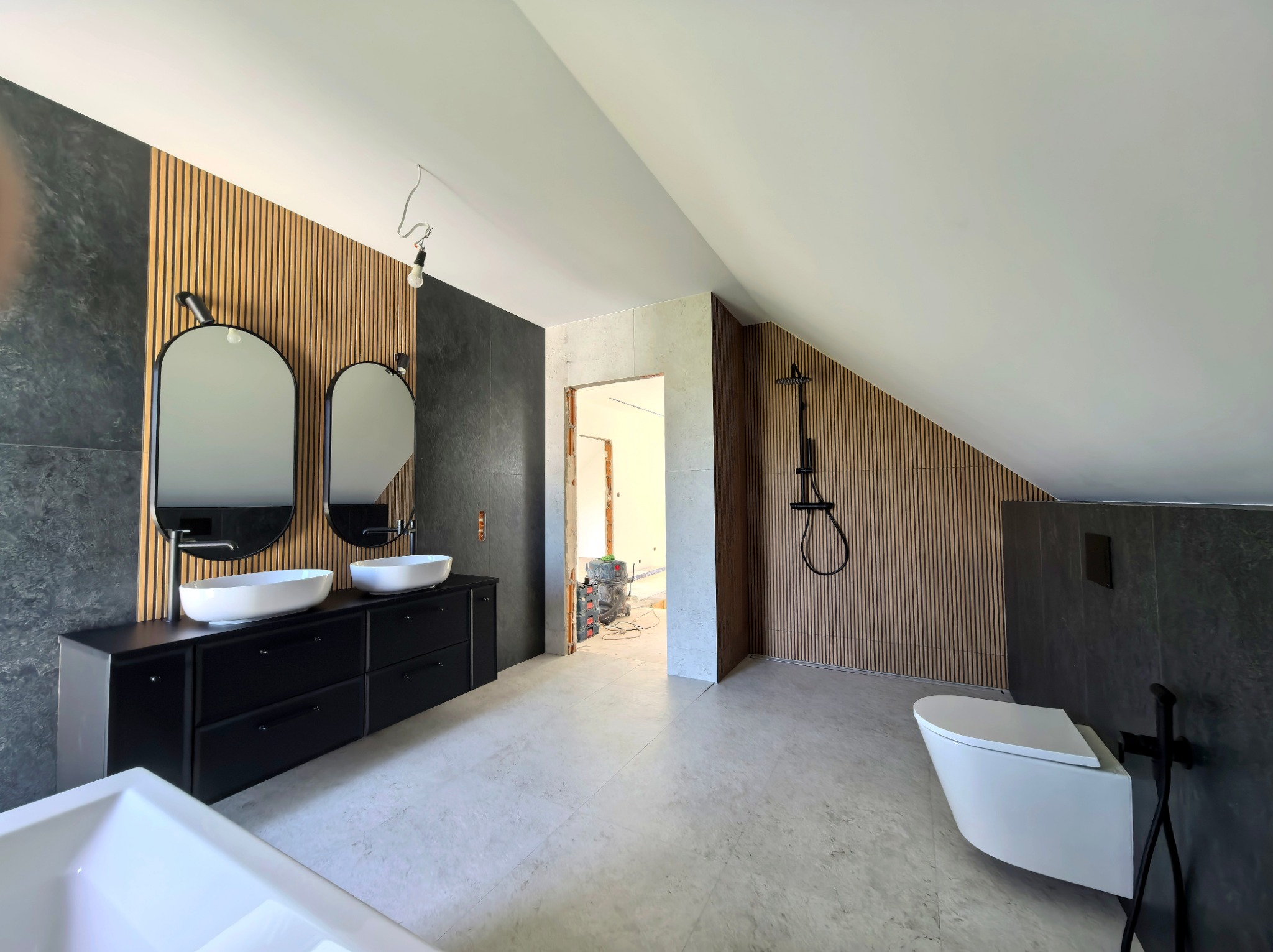 Nowoczesna łazienka na poddaszu z drewnianymi panelami ściennymi, czarną armaturą i umywalkami nablatowymi. Widoczna wanna i toaleta w minimalistycznym stylu.