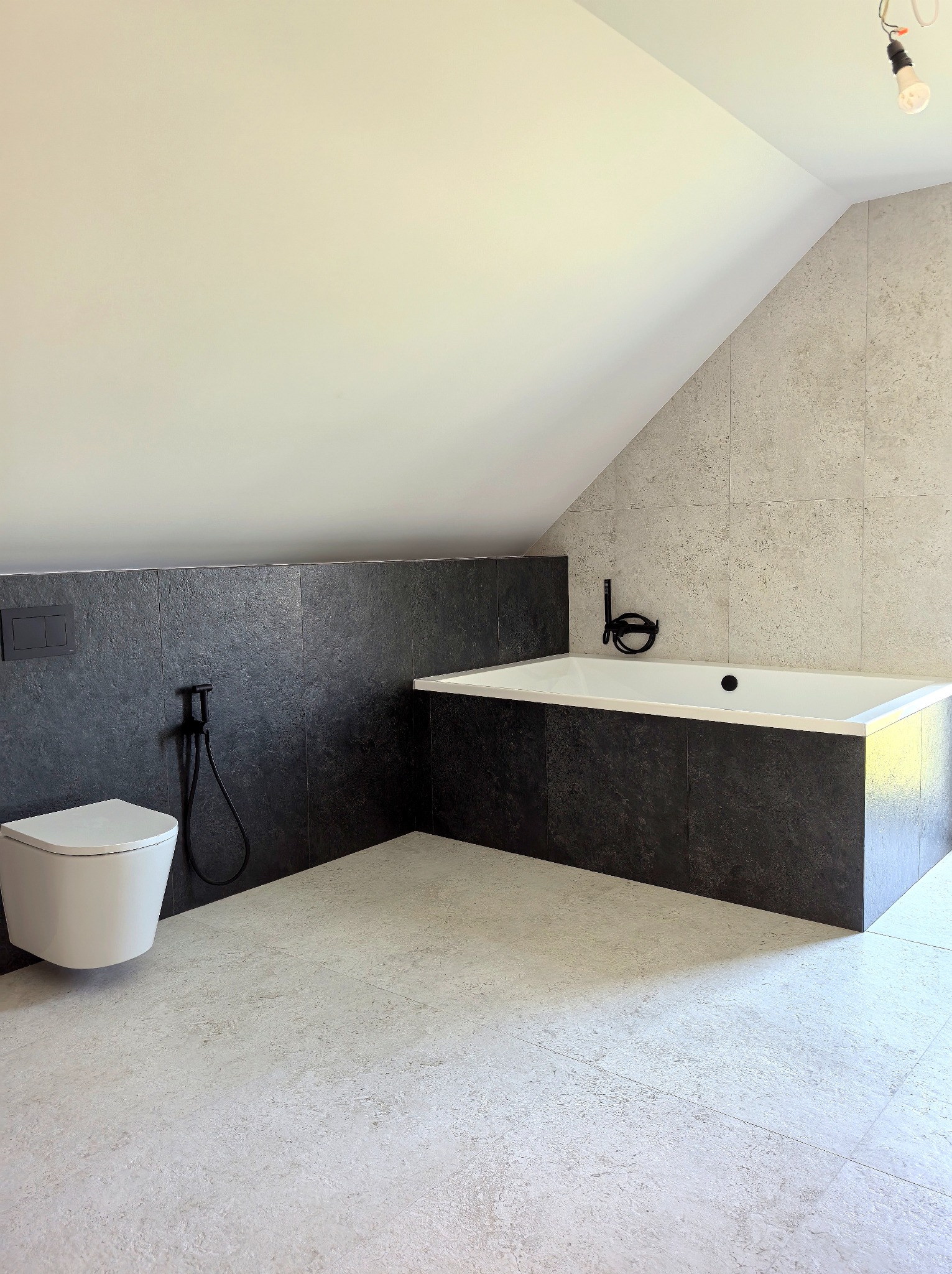 Nowoczesna łazienka na poddaszu z wanną obudowaną ciemnymi płytkami i podwieszaną toaletą. Jasne płytki na podłodze i ścianie tworzą kontrast.