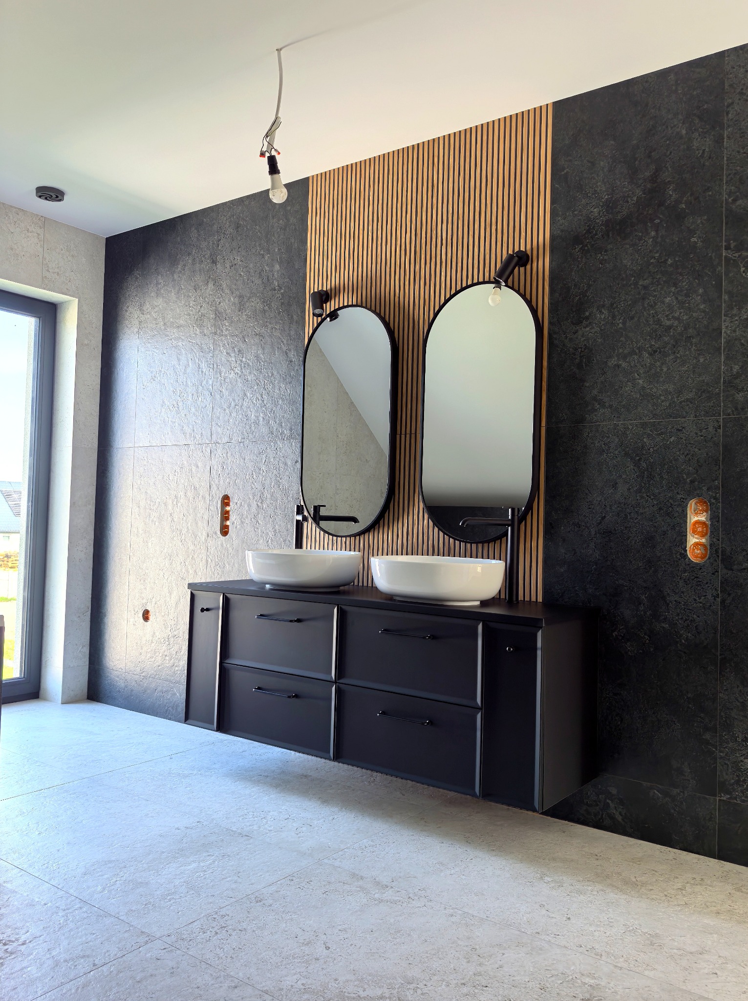 Nowoczesna łazienka z dwoma umywalkami nablatowymi, czarną szafką, lustrami w owalnych ramach i akcentem drewnianych lameli na ścianie. Widoczne płytki imitujące kamień.