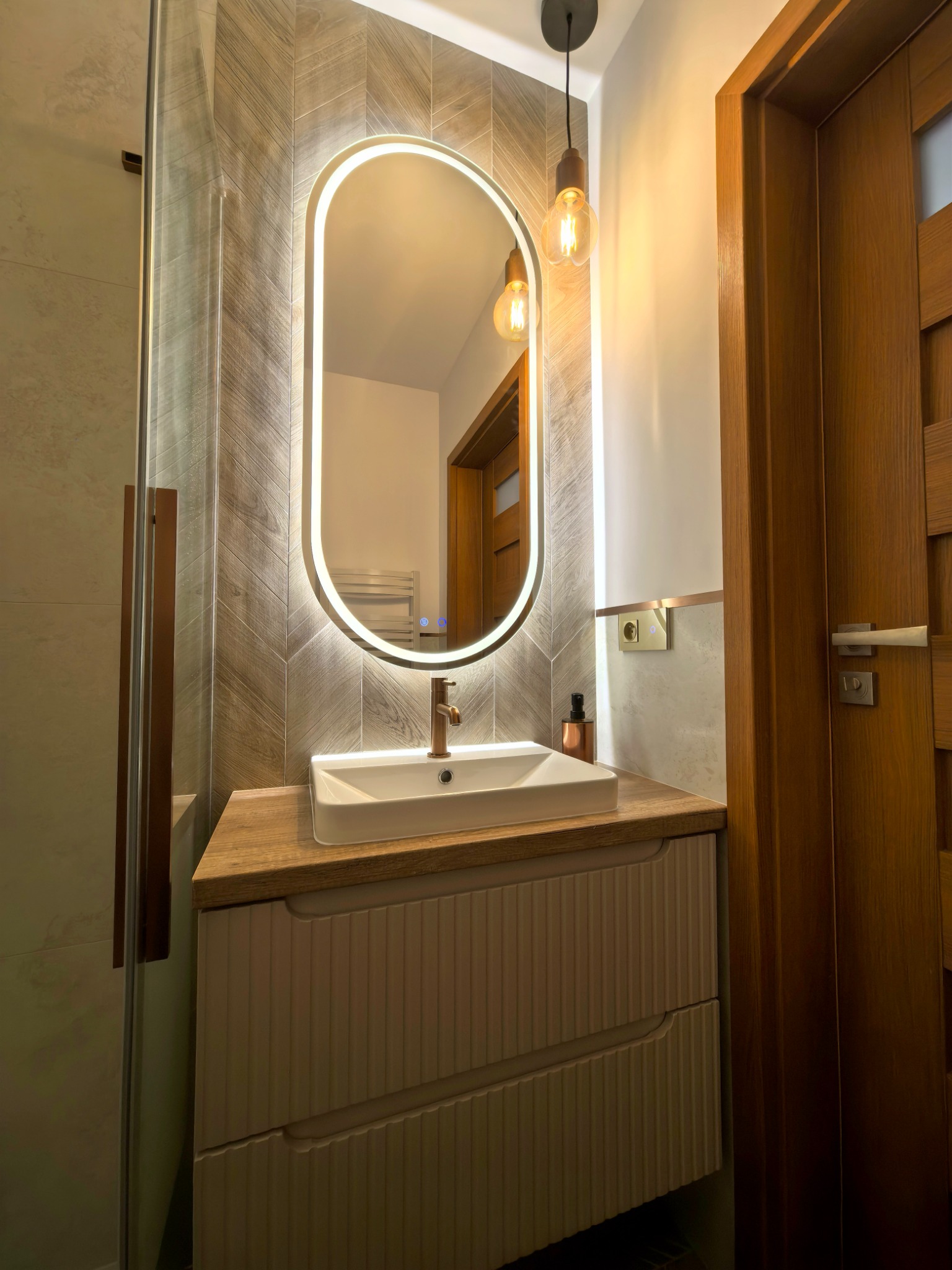 Nowoczesna łazienka z umywalką nablatową, podświetlanym lustrem w kształcie owalu i drewnianymi akcentami. Ściana wyłożona płytkami imitującymi drewno w jodełkę.