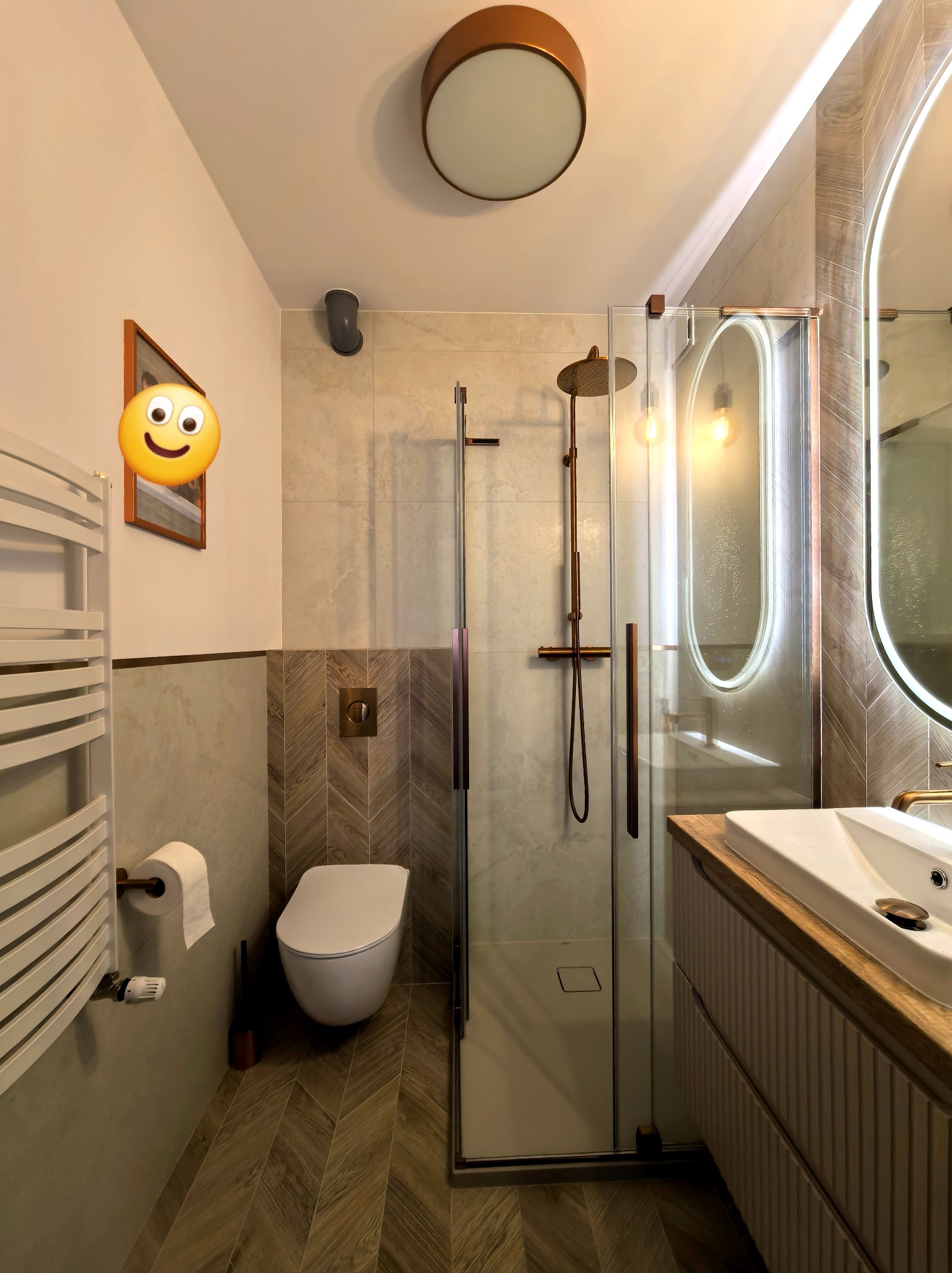 Nowoczesna łazienka z kabiną prysznicową typu walk-in, umywalką na szafce z drewnianym blatem i podświetlanym lustrem. Uwagę zwracają płytki ułożone w jodełkę na podłodze i ścianie.