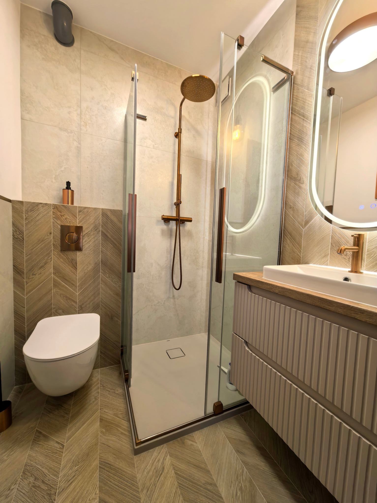 Nowoczesna łazienka z kabiną prysznicową typu walk-in, umywalką na szafce z frezowanymi frontami i podświetlanym lustrem. Uwagę zwracają płytki ułożone w jodełkę.