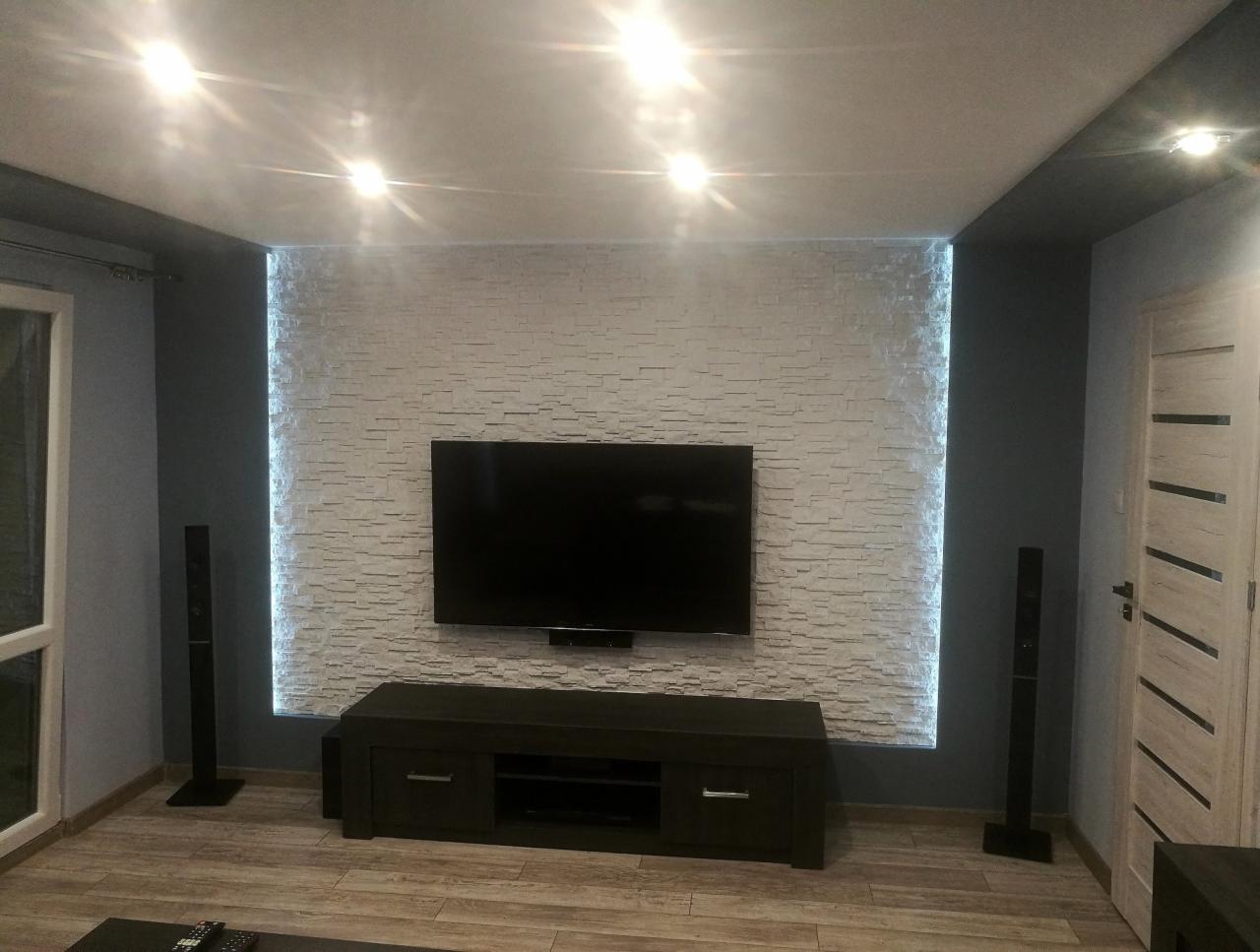 Salon z dekoracyjną ścianą z białej cegły, podświetloną ledami, na której wisi telewizor. Pod telewizorem ciemna szafka RTV, a po bokach głośniki. Widoczna drewnopodobna podłoga i drzwi z poziomymi...