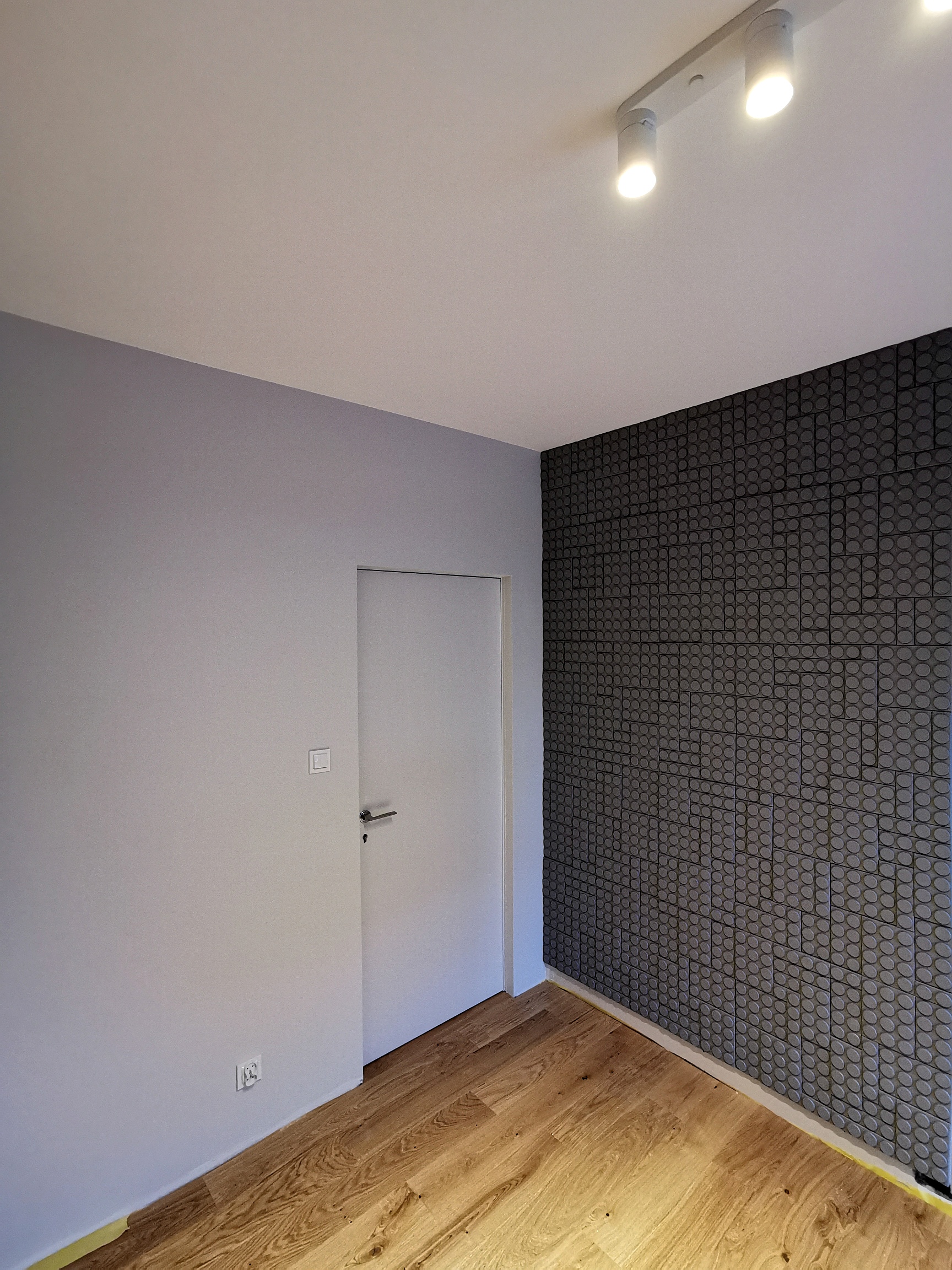Wnętrze pokoju po remoncie: ściana pomalowana na szaro, fragment ściany wykończony dekoracyjnymi panelami z motywem okręgów, drewniana podłoga, białe drzwi i nowoczesne oświetlenie sufitowe.