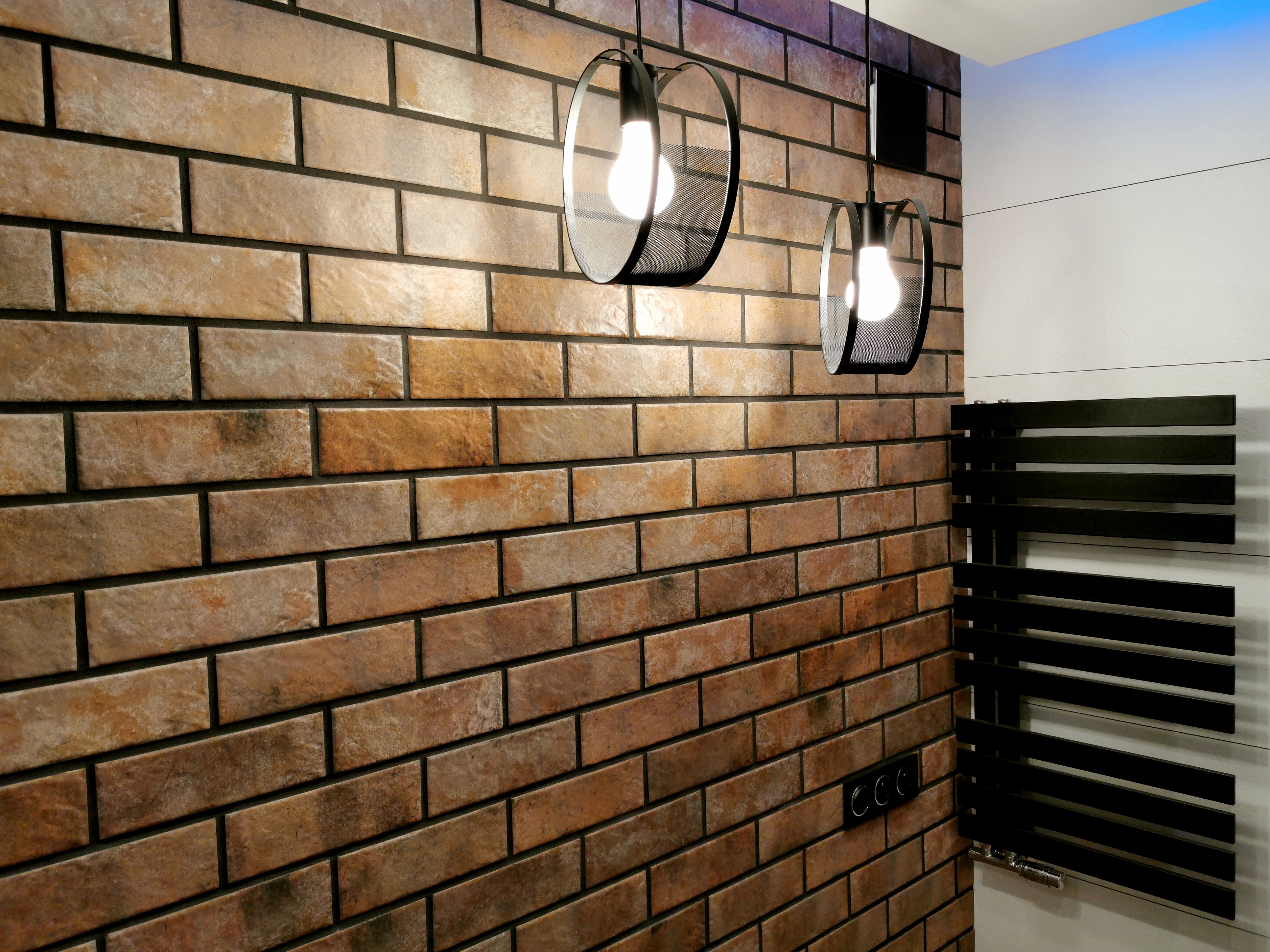 Wykończona ściana z cegiełek w łazience, oświetlona dwoma wiszącymi lampami z czarnymi, ażurowymi kloszami, obok grzejnik drabinkowy w kolorze czarnym.