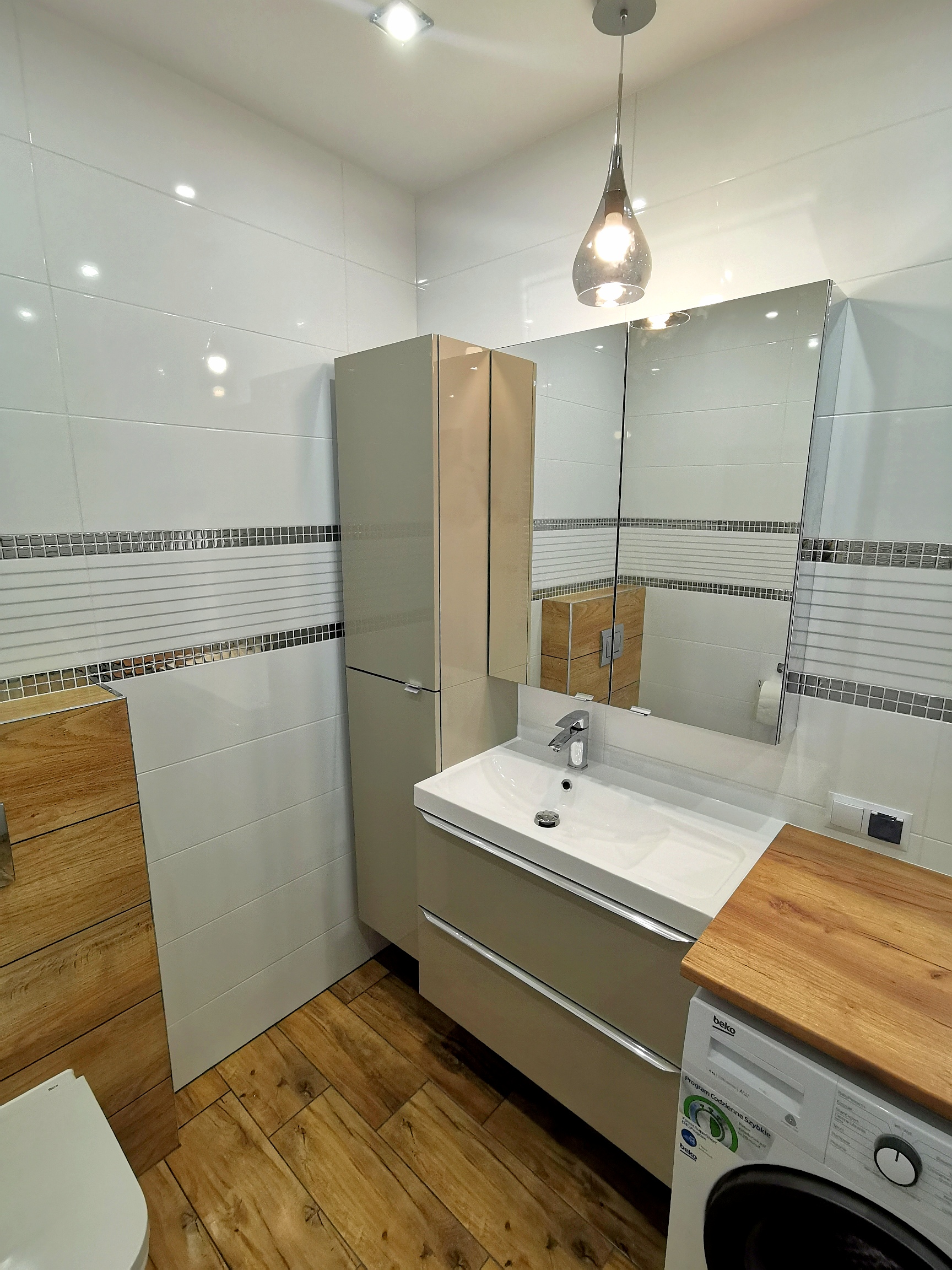 Nowoczesna łazienka z białymi płytkami, drewnianymi akcentami i wiszącą lampą w kształcie kropli, pralka z drewnianym blatem obok szafki umywalkowej.