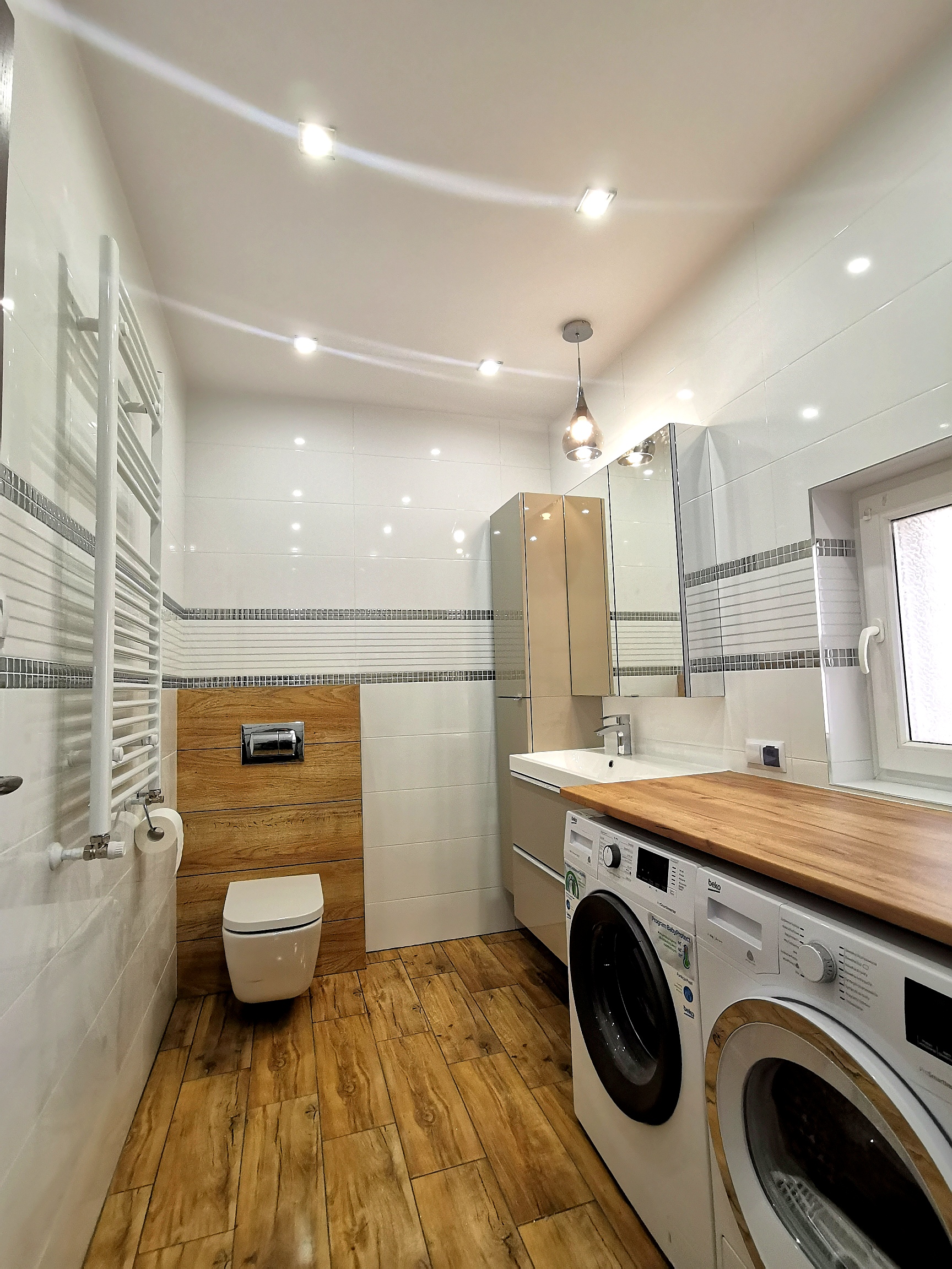 Nowoczesna łazienka z drewnianymi akcentami: płytki imitujące drewno na podłodze i obudowie toalety, białe płytki na ścianach z dekoracyjnym pasem, podwieszana toaleta, umywalka z szafką, lustro...