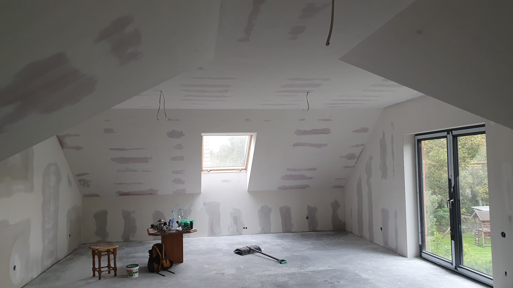 Poddasze w trakcie prac wykończeniowych: ściany i sufit z widocznymi plamami po szpachlowaniu, przygotowane do dalszych prac malarskich, widoczne narzędzia i materiały budowlane, okno dachowe...