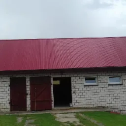 Budynek gospodarczy z czerwonym dachem z blachy trapezowej, białe ściany z cegły silikatowej, drewniane wrota, pochmurne niebo.