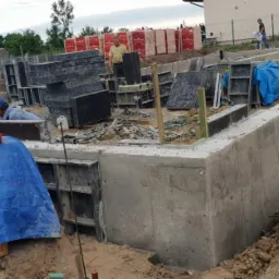 Wylane fundamenty domu z widocznymi szalunkami, przykryte folią zabezpieczającą, na tle placu budowy z cegłami i pracującymi osobami.