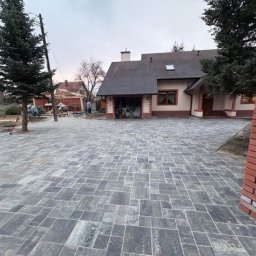 MJD MICHAŁ DACZ - Ułożona szara kostka brukowa na podjeździe domu jednorodzinnego, w tle widoczne prace budowlane przy drewnianej konstrukcji, częściowo ukończony murek z czerwonej cegły w prawym dolnym rogu.