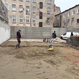 MJD MICHAŁ DACZ - Plac budowy z częściowo ułożoną kostką, zagęszczarką i pracownikami na tle miejskiej zabudowy.