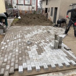 MJD MICHAŁ DACZ - Trzech mężczyzn układa szarą kostkę brukową na piasku, widoczny stos kostki, gumowy młotek i ubijak, w tle budynek i pryzma ziemi.