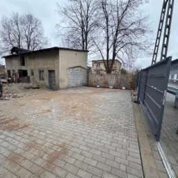 Szara kostka brukowa na podw&oacute;rku z widocznym, częściowo zamontowanym, przesuwnym ogrodzeniem metalowym w kolorze antracytowym oraz starym budynkiem gospodarczym w tle.
