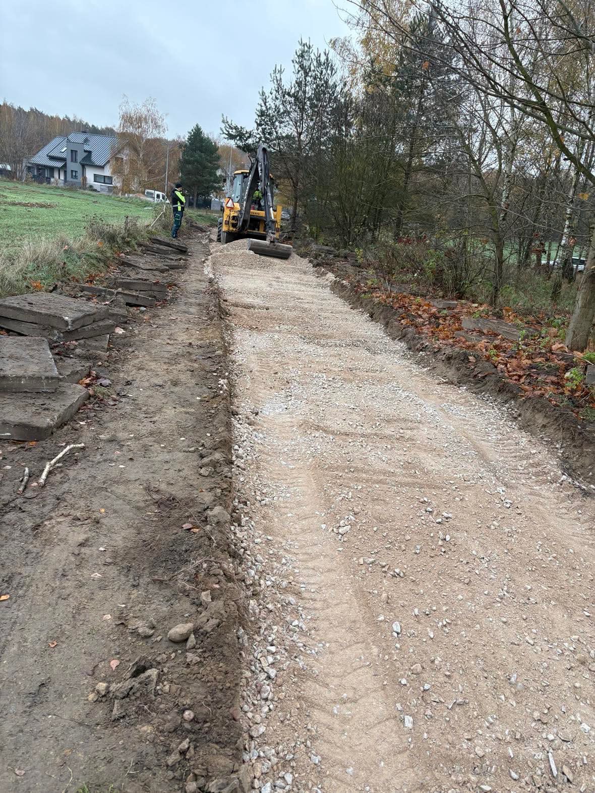 Przygotowanie podłoża pod nawierzchnię: wyrównywanie terenu kruszywem za pomocą walca wibracyjnego, widoczny pracownik w odblaskowej kamizelce i fragmenty betonowych płyt.