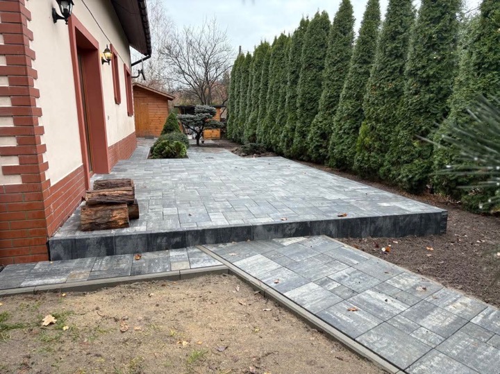 Szara, prostokątna nawierzchnia z kostki brukowej przed domem, z ułożonymi schodami i ścieżką, obok ułożone drewno, w tle rząd wysokich, zielonych tuj.