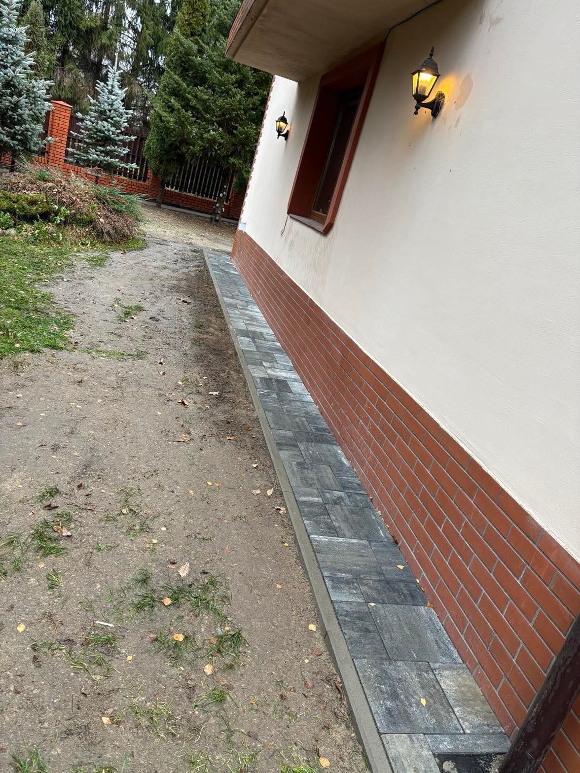 Ułożona szara kostka brukowa wzdłuż ściany budynku z ceglaną podmurówką i oświetleniem zewnętrznym, widoczne elementy ogrodu w tle.