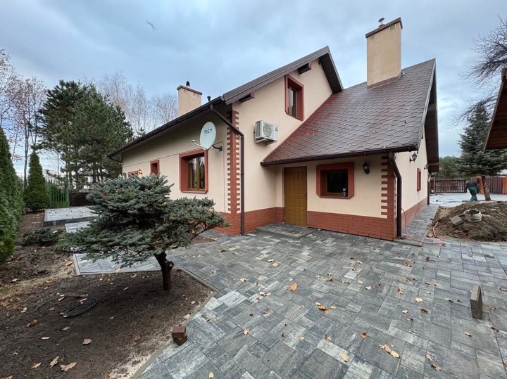 Ułożona kostka brukowa w odcieniach szarości przed domem z beżową elewacją i brązowymi akcentami, widoczny fragment ogrodu z młodym świerkiem i przygotowane podłoże pod dalsze prace.