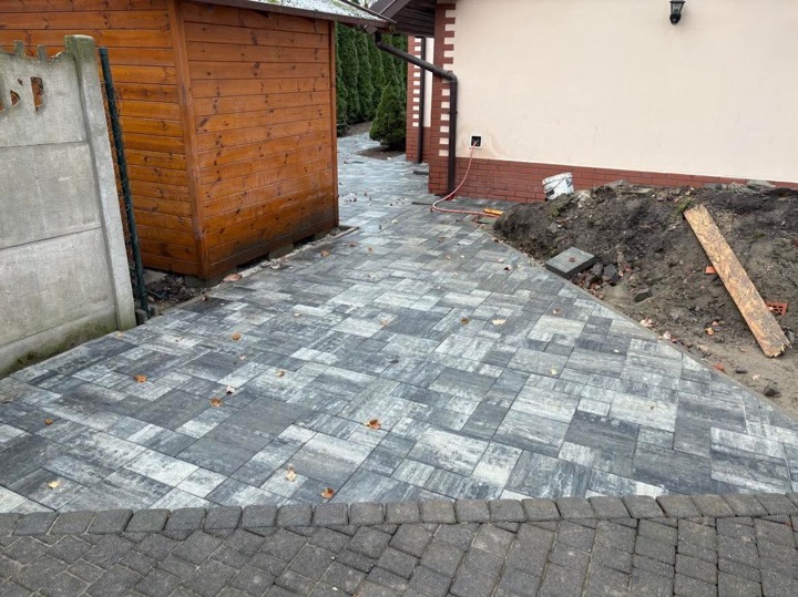 Ułożona szara kostka brukowa w geometryczny wzór prowadząca od drewnianej altany wzdłuż budynku, widoczne ślady prac ziemnych i pojedyncze opadłe liście.