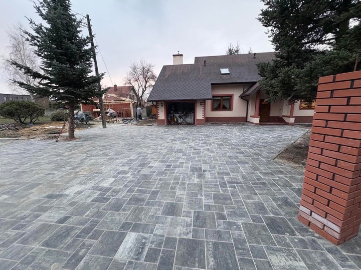 Ułożona szara kostka brukowa na podjeździe domu jednorodzinnego, w tle widoczne prace budowlane przy drewnianej konstrukcji, częściowo ukończony murek z czerwonej cegły w prawym dolnym rogu.