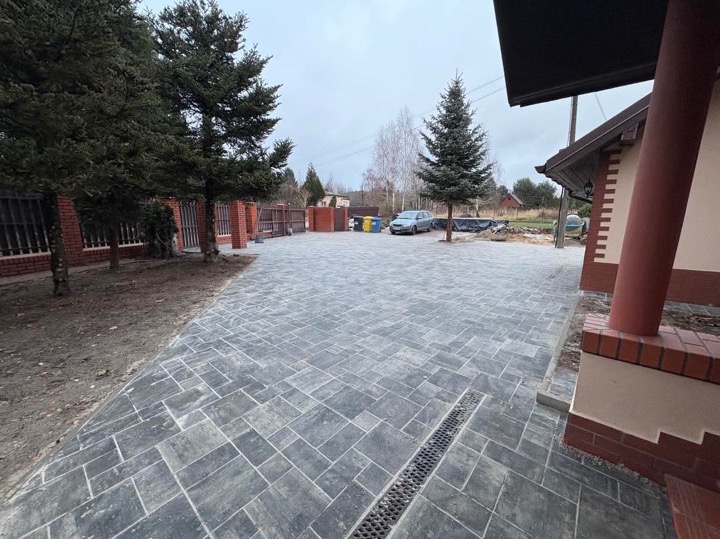 Ułożona szara kostka brukowa na podjeździe domu jednorodzinnego z odpływem liniowym, widok na ogrodzenie z czerwonej cegły i drzewa iglaste w pochmurny dzień.