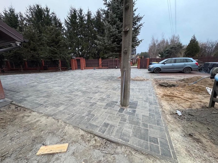 Ułożona szara kostka brukowa na podjeździe z drzewem wyrastającym pośrodku, widoczne ogrodzenie z ceglanymi słupkami i stojący samochód.