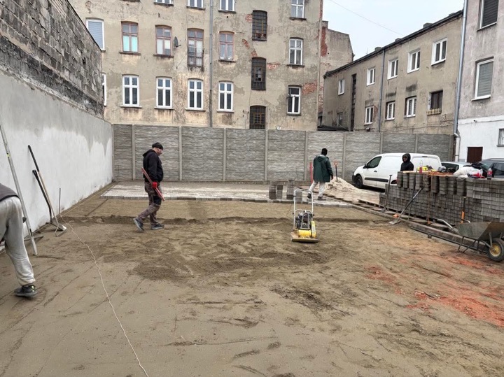 Plac budowy z częściowo ułożoną kostką, zagęszczarką i pracownikami na tle miejskiej zabudowy.