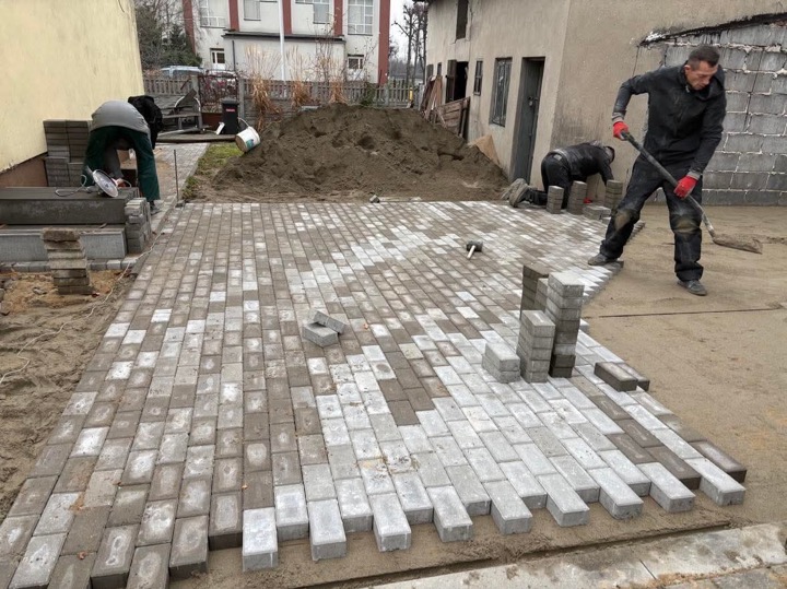 Trzech mężczyzn układa szarą kostkę brukową na piasku, widoczny stos kostki, gumowy młotek i ubijak, w tle budynek i pryzma ziemi.