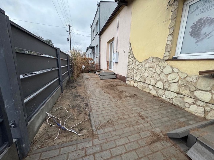 Nowoczesne, grafitowe ogrodzenie panelowe, ułożona szara kostka brukowa wzdłuż posesji, z widocznymi przewodami elektrycznymi w piasku i fragmentem elewacji budynku z oknem salonu fryzjerskiego w tle.