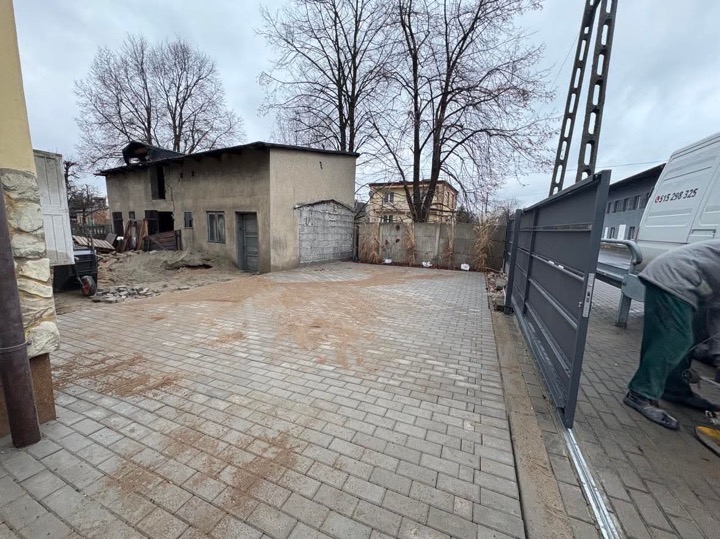 Szara kostka brukowa na podwórku z widocznym, częściowo zamontowanym, przesuwnym ogrodzeniem metalowym w kolorze antracytowym oraz starym budynkiem gospodarczym w tle.