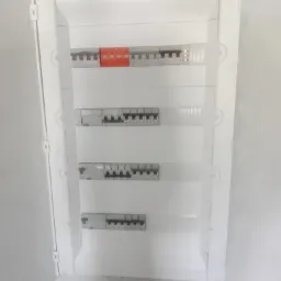 Otwarta, biała skrzynka rozdzielcza z widocznymi bezpiecznikami i wyłącznikami różnicowoprądowymi na białej ścianie.