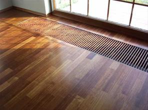 Parkiet z drewna egzotycznego ułożony w jodełkę francuską, przy oknie z wbudowanym w podłogę grzejnikiem kanałowym z czarną kratką.