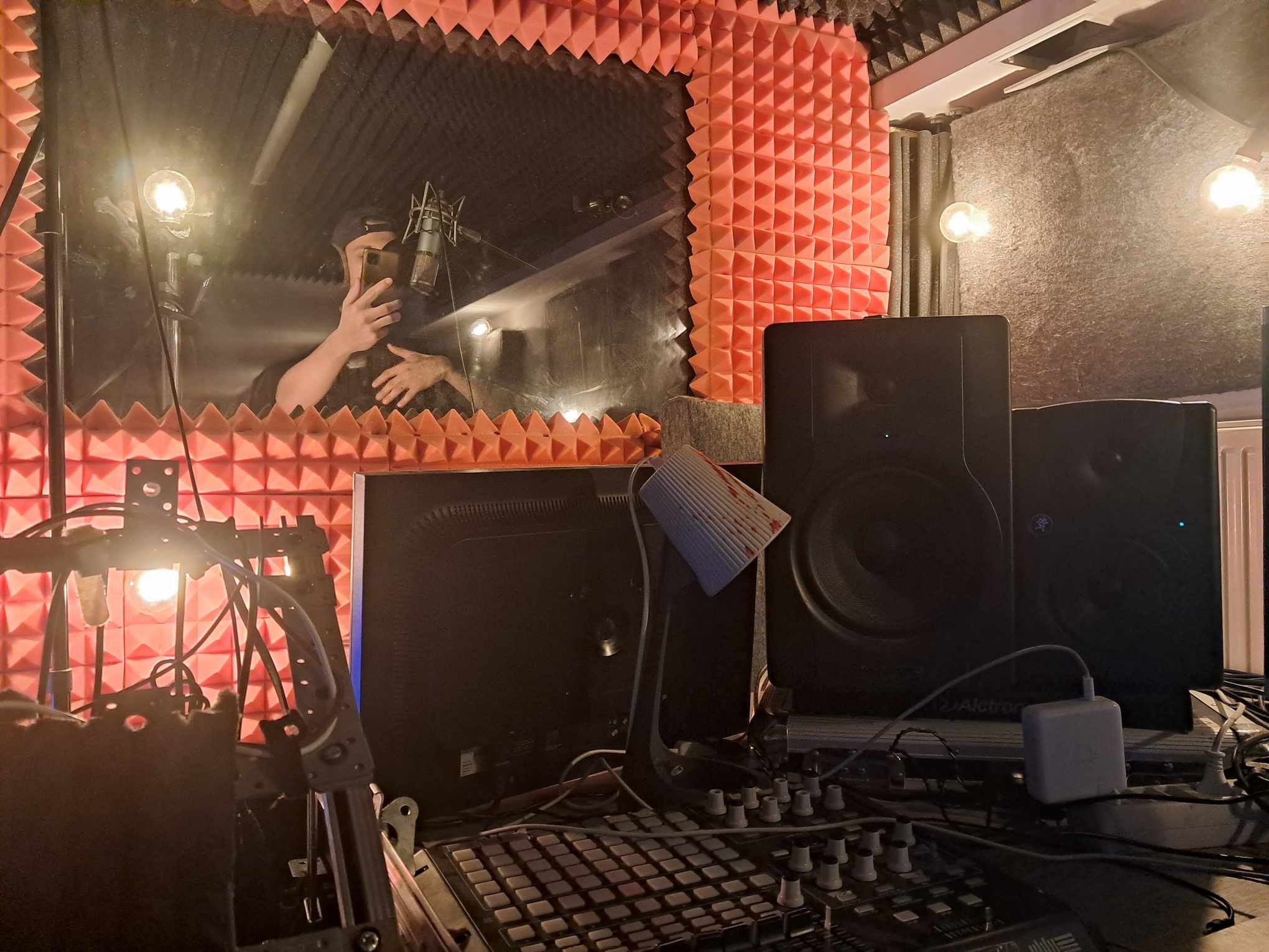 Osoba robi selfie w kabinie studia nagrań z mikrofonem i panelami akustycznymi, widoczne monitory studyjne i mikser. Scena w ciepłym oświetleniu.