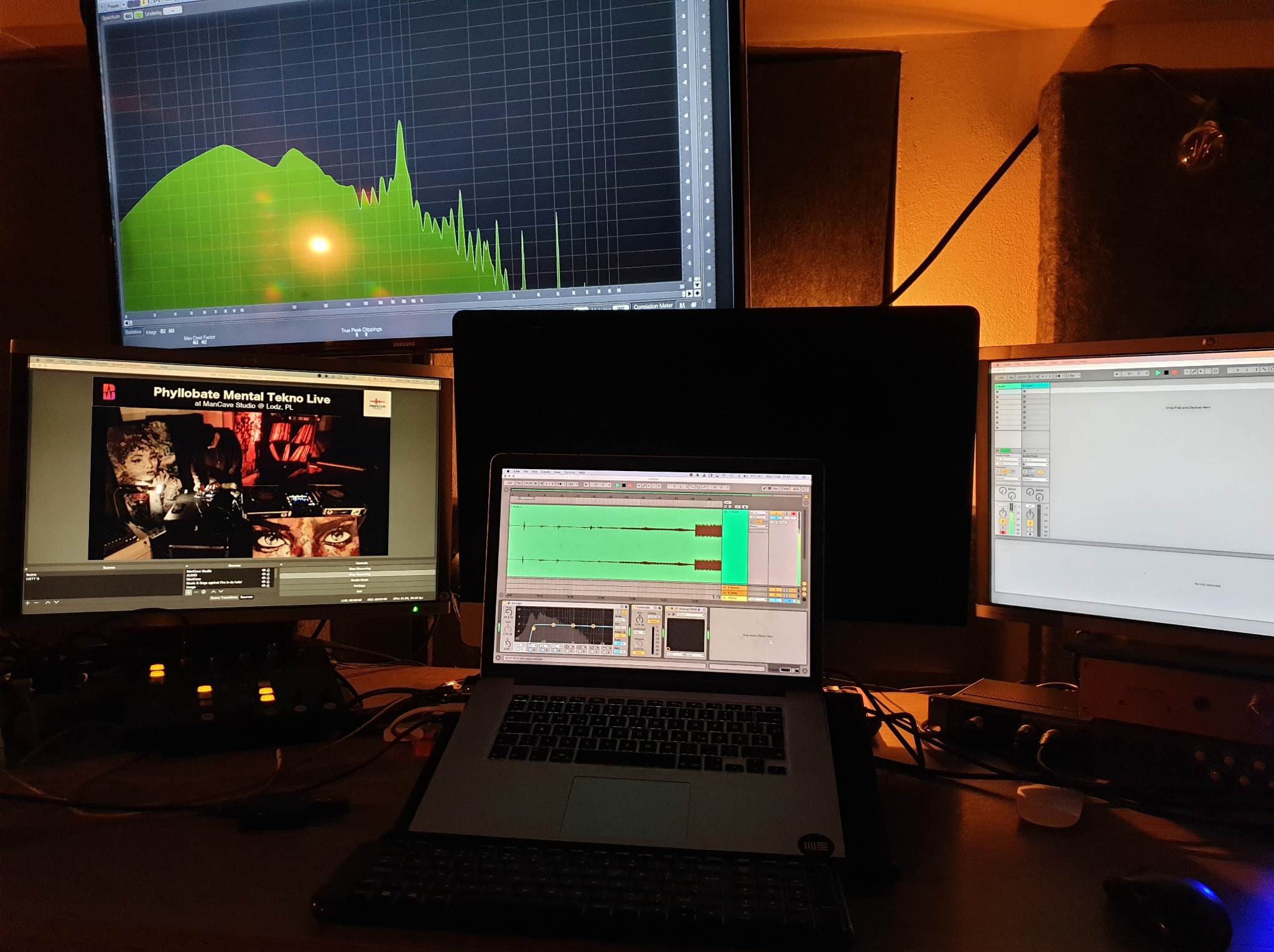 Stanowisko pracy w studio nagrań: monitory z oprogramowaniem audio, laptop z edytorem dźwięku. Widoczne analizatory spektrum i interfejs DAW. Ciepłe oświetlenie.