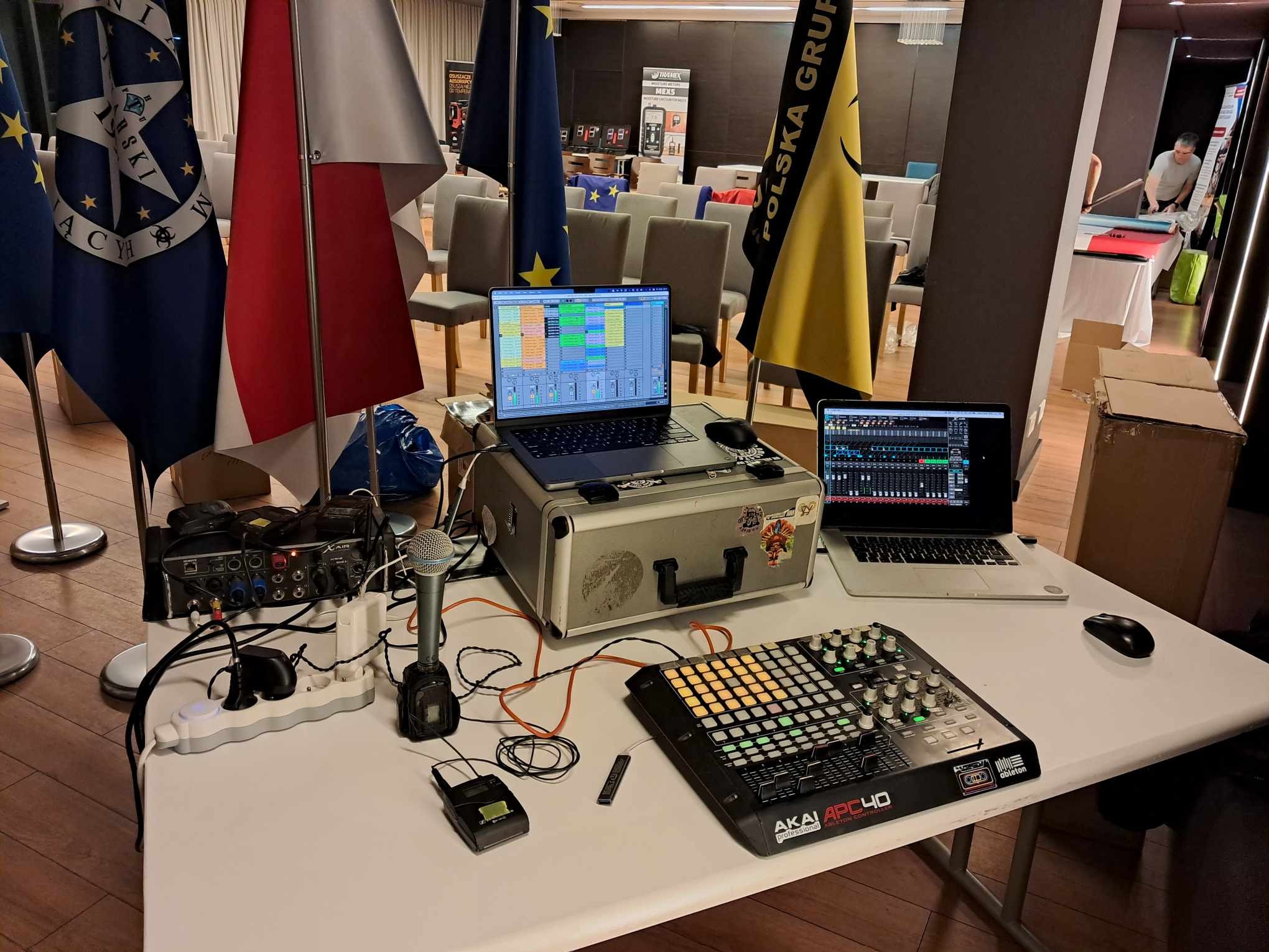 Stanowisko DJ-a z laptopami, mikserem Akai APC40 i mikrofonem na stole, w tle flagi i krzesła. Sprzęt gotowy do występu.
