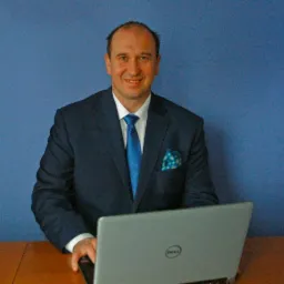 Uśmiechnięty mężczyzna w garniturze i krawacie, siedzący za biurkiem z laptopem Dell na tle niebieskiej ściany. Widoczna poszetka w kieszonce.