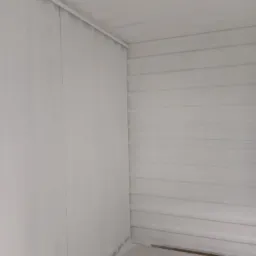 Wnętrze pomalowanego na biało pomieszczenia z blachy falistej, widoczne łączenia blach i fragmenty drewnianych elementów na podłodze.