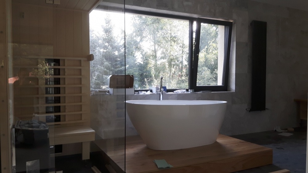 Nowoczesna łazienka z wolnostojącą białą wanną na drewnianym podeście, widok na zewnątrz przez duże okno, obok sauna z kamieniami wewnątrz, minimalistyczny grzejnik na ścianie.