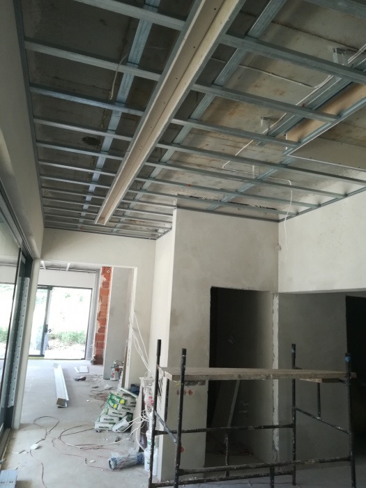 Metalowa konstrukcja sufitu podwieszanego w trakcie montażu, widoczne przewody, rusztowanie i worki z zaprawą w surowym wnętrzu.