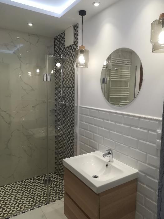 Nowoczesna łazienka po remoncie: kabina prysznicowa z marmurowymi płytkami, umywalka z drewnianą szafką, okrągłe lustro i wisząca lampa z szarym kloszem.