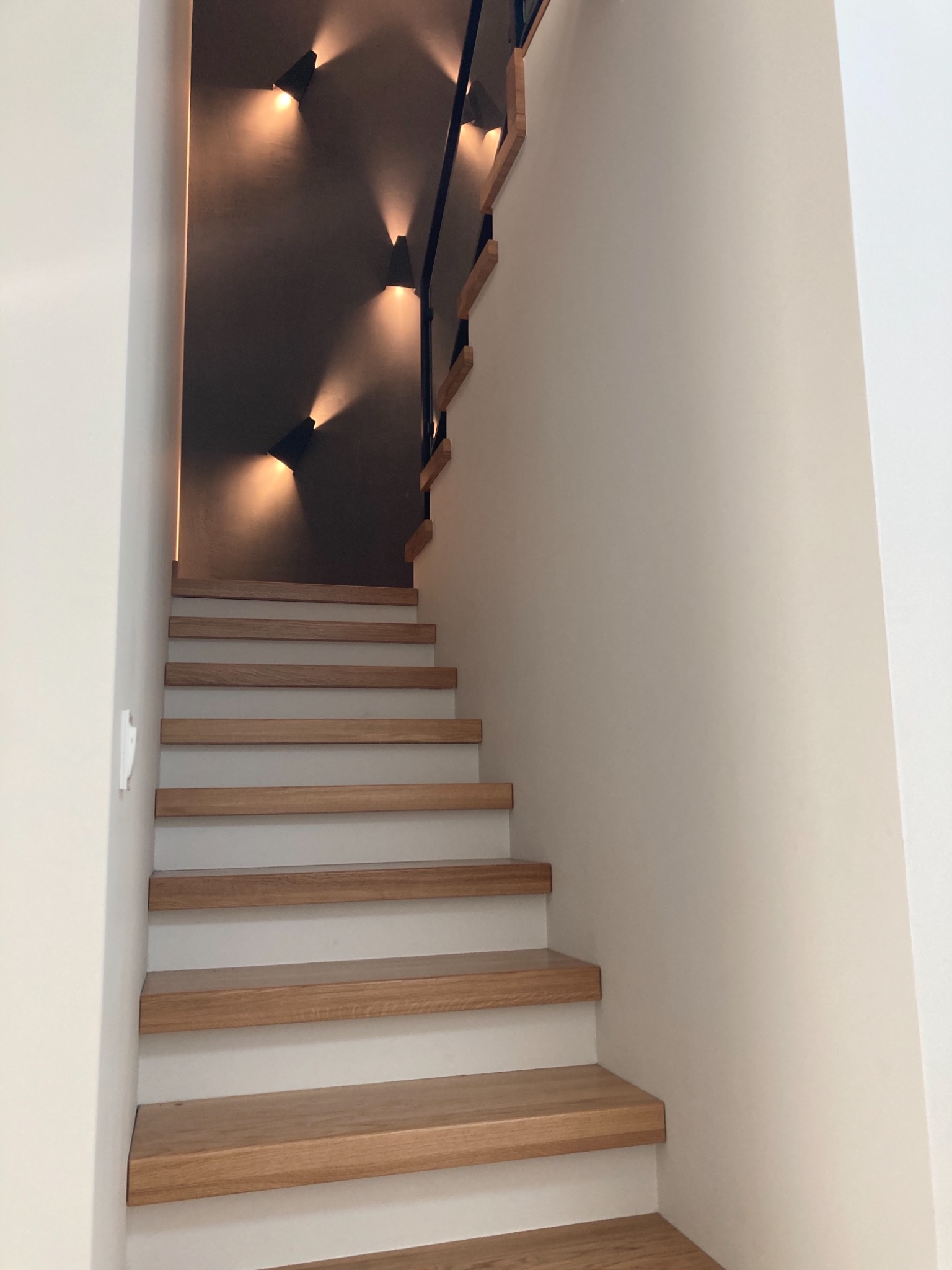 Drewniane schody z białymi podstopnicami, oświetlone trójkątnymi lampami ściennymi, czarna balustrada