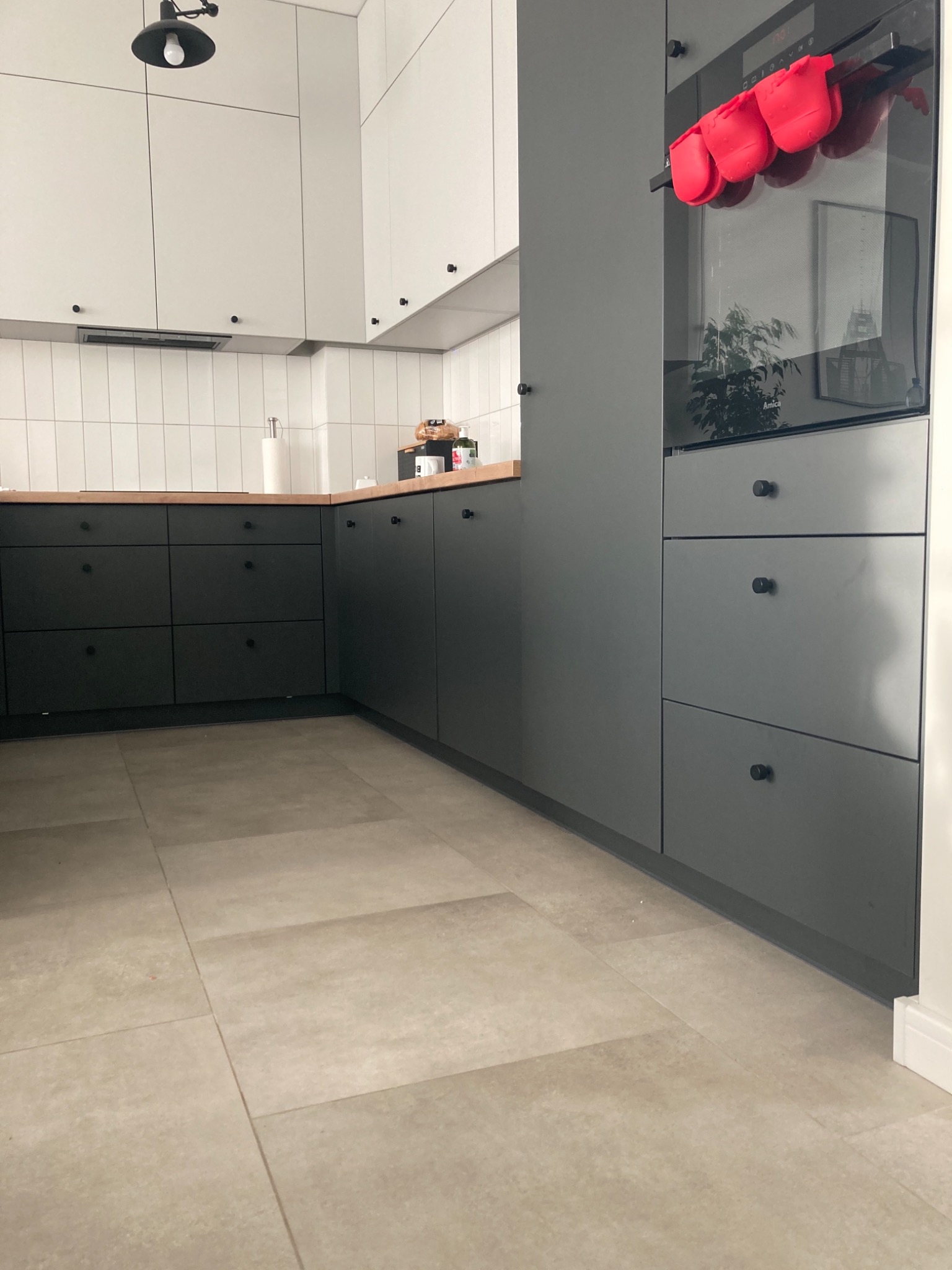 Nowoczesna kuchnia z szarymi szafkami z okrągłymi uchwytami, drewnianym blatem i białymi płytkami na ścianie. Na piekarniku wiszą czerwone rękawice kuchenne.