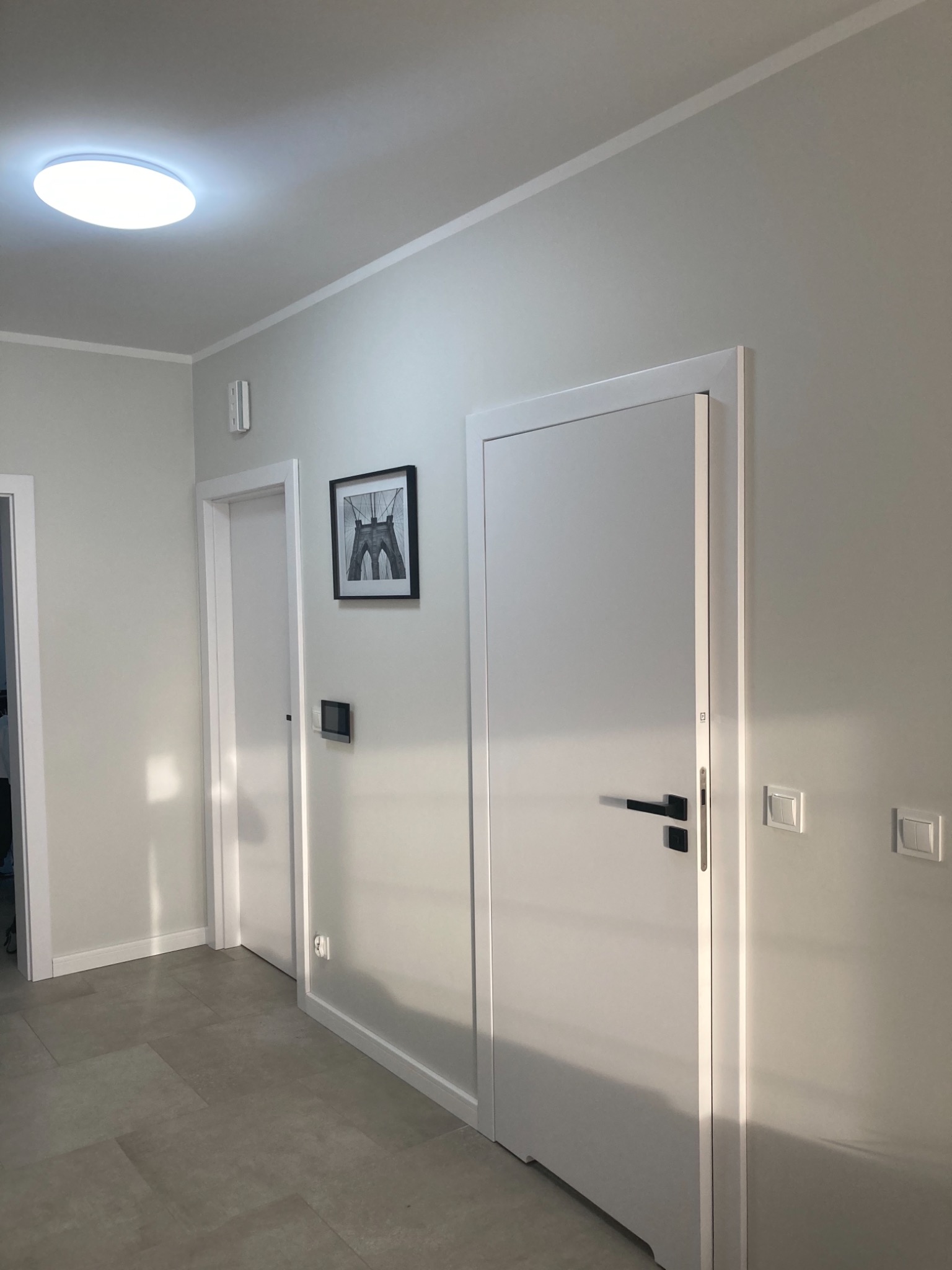Jasne wnętrze korytarza z dwiema białymi drzwiami, szarą podłogą z płytek i obrazem w czarnej ramie na ścianie, oświetlone okrągłą lampą sufitową LED.