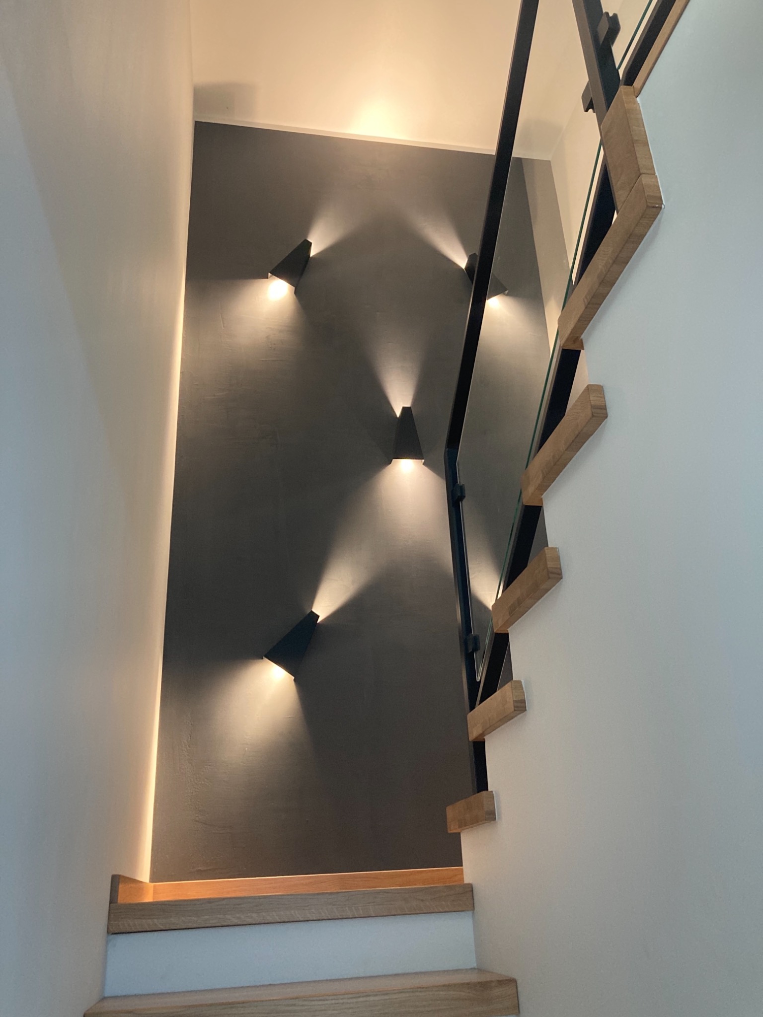 Nowoczesna klatka schodowa z drewnianymi stopniami, czarną balustradą ze szklanym wypełnieniem i szarą ścianą oświetloną przez trzy trójkątne kinkiety.