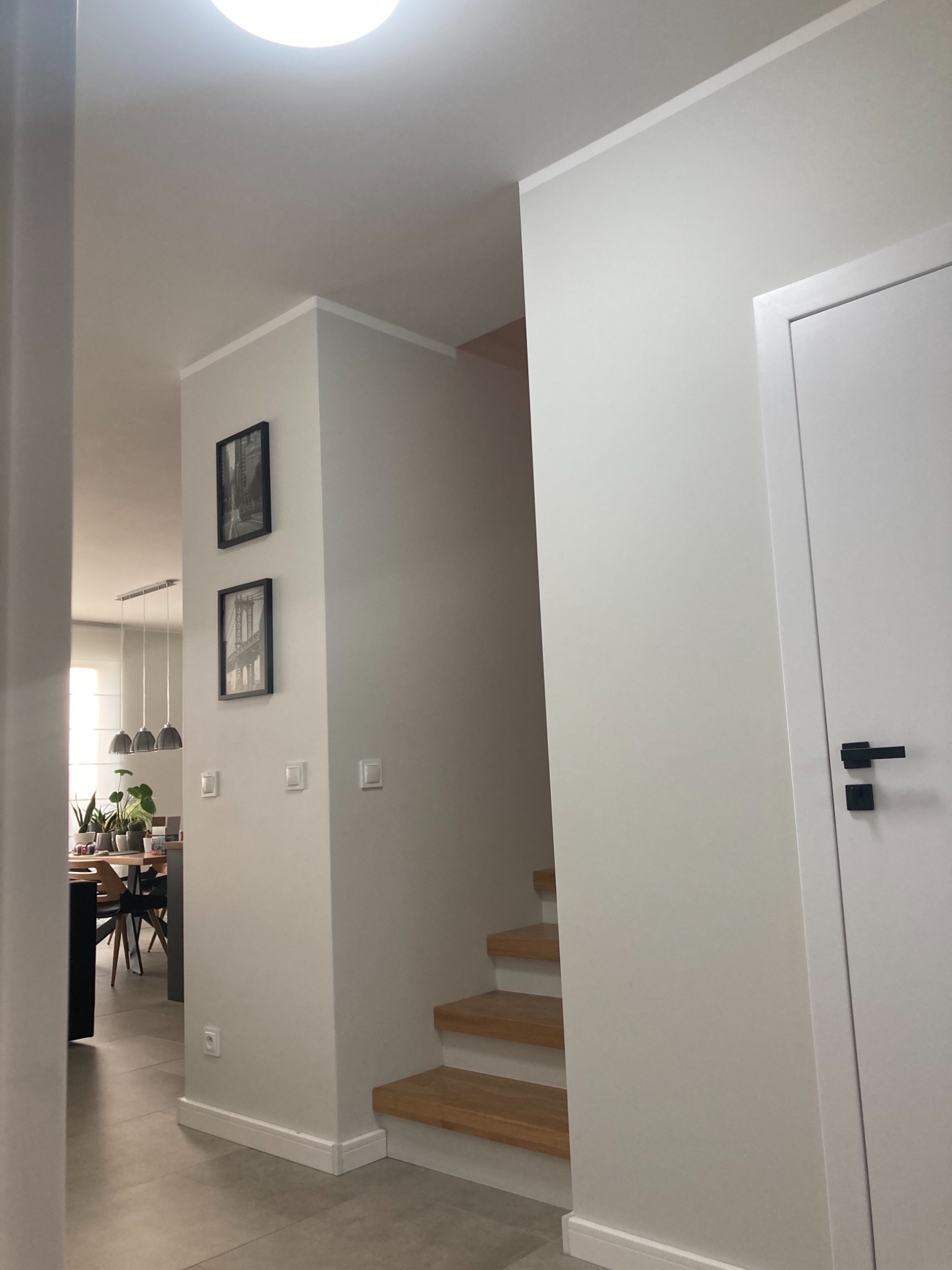 Jasne wnętrze domu po remoncie, widoczne schody z drewnianymi stopniami, białe ściany z dwoma czarno-białymi obrazami w ramkach, drzwi z czarną klamką oraz fragment jadalni z roślinami.