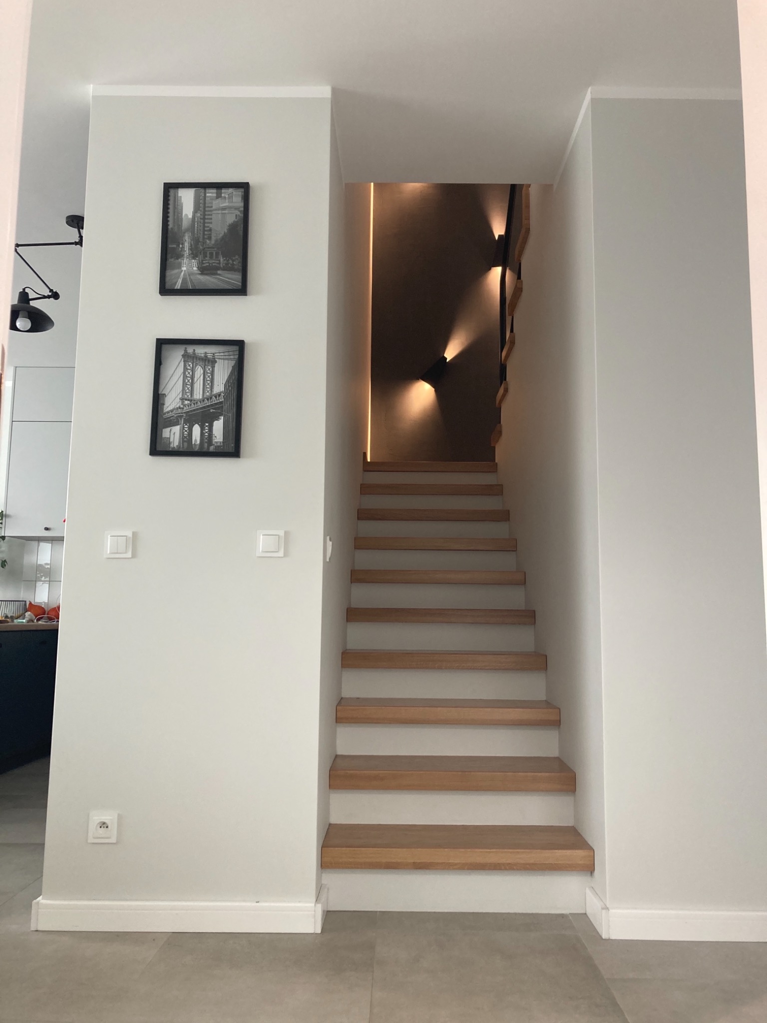 Nowoczesne schody drewniane z podświetleniem LED w minimalistycznym wnętrzu, na ścianie czarno-białe zdjęcia w ramkach.