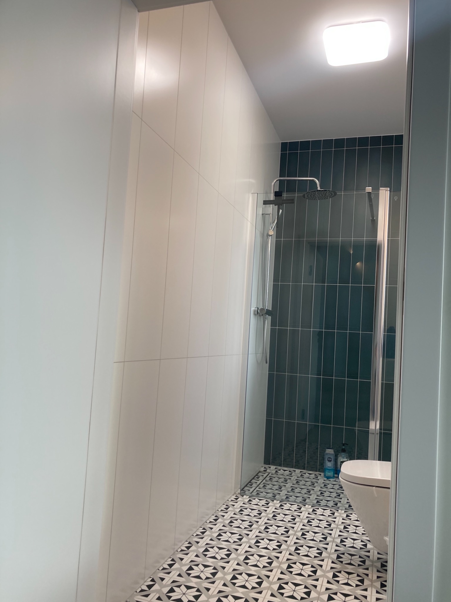 Wykończona łazienka z kabiną prysznicową za szkłem, pionowe ciemnozielone płytki na ścianie, wzorzysta czarno-biała podłoga i minimalistyczna toaleta.