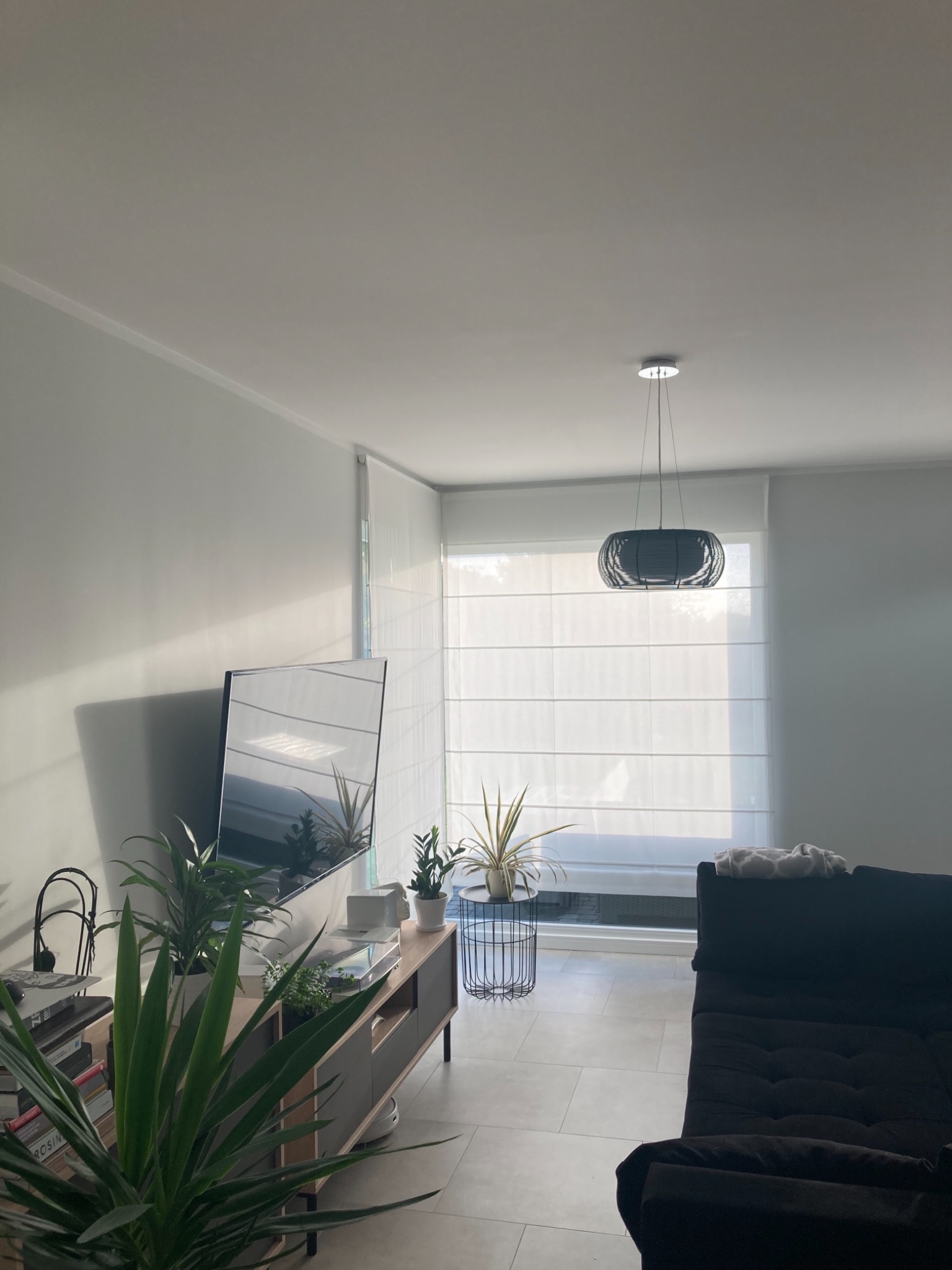 Jasne wnętrze salonu z szarymi płytkami podłogowymi, zasłoniętym oknem roletami rzymskimi, ciemną sofą i niską szafką RTV z roślinami doniczkowymi, oświetlone naturalnym światłem.