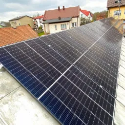 Panele fotowoltaiczne zamontowane na płaskim dachu budynku mieszkalnego, widok z góry, w tle zabudowa jednorodzinna i pochmurne niebo.