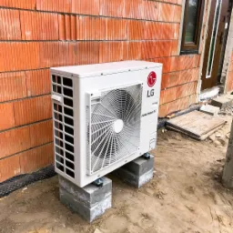 Biała zewnętrzna jednostka klimatyzacji LG Therma V zamontowana na betonowych bloczkach przed budynkiem z czerwonej cegły dziurawki w trakcie budowy, z widocznym oknem i drzwiami.